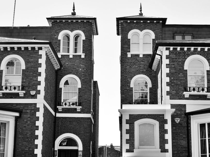 A black and white photograph of a Victorian-era residential building showing two contrasting brick towers with decorative white trim around arched and rectangular windows. The towers are connected by 